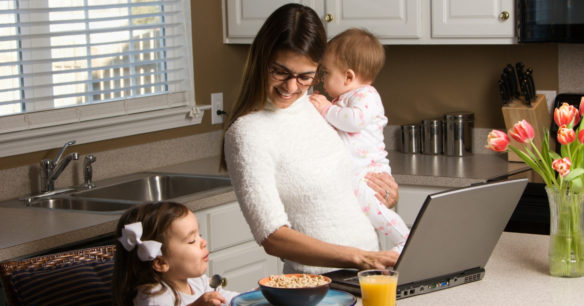 woman multitasking on computer holding a baby and feeding a toddler